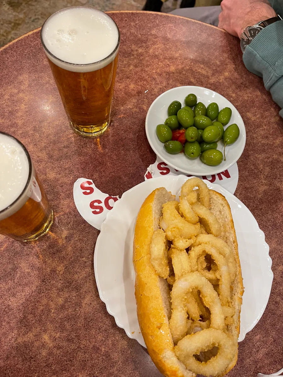 sandwich with fried calamari inside it, a small plate of olives on the side and two beers.