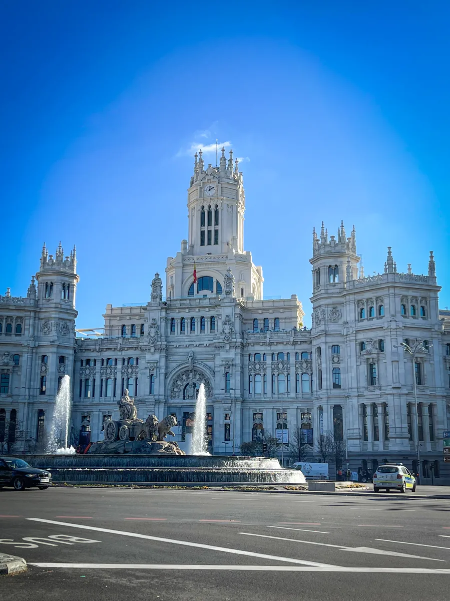 front of a palace-like building with a central tower in the middle and many smaller turrets along the left and right side of the building with a large water fountain at the front with a horse and carriage inside the fountain.
