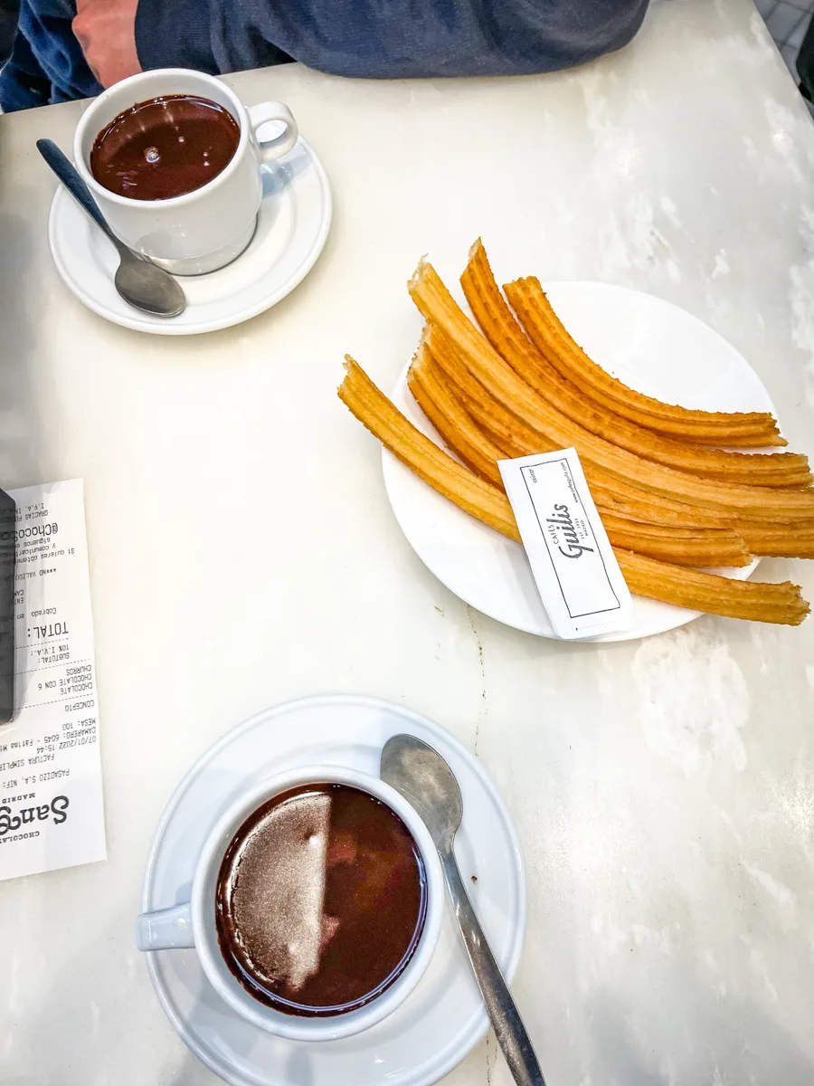 churros stacked on a place with two mugs of rich thick hot chocolate on either side of the table with spoons next to the hot chocolate.